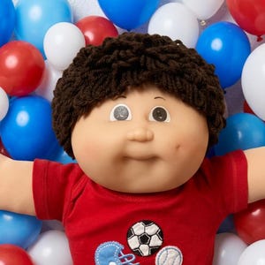 May include: A doll with dark brown yarn hair and a round face wearing a red t-shirt with embroidered sports ball and helmet patches, paired with black pants and white shoes, surrounded by colorful balloons and various small toy vehicles and balls.