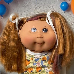 May include: A doll with brown hair in pigtails, wearing a yellow floral top and white shoes, surrounded by orange, blue, and white balloons, several plush teddy bears in orange and blue, and various colorful wrapped candies and lollipops.