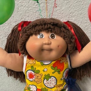 May include: A doll with brown yarn hair in pigtails, wearing a yellow sleeveless top with a strawberry and bear print, white ruffled socks, and white shoes, surrounded by red, green, and yellow balloons and four small plush bears.