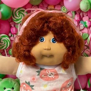May include: A doll with curly auburn hair and blue eyes wearing a sleeveless white top with a pink strawberry and bunny print, white socks, and white shoes, surrounded by pink and green balloons, teddy bears, and various candy pieces.
