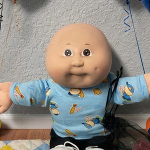 May include: A bald doll with a light skin tone wearing a light blue long-sleeve shirt with a baseball print and black pants. The doll is surrounded by two balloon bouquets in blue, black, and beige, toy trains, cars, a soccer ball, and cookies.