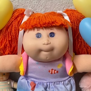 May include: A doll with bright orange yarn hair in pigtails, wearing a light purple dress with a fish print, pink shoes, and white lace socks. The doll is surrounded by heart-shaped balloons, a pink teddy bear, and a plate of chocolate brownies.