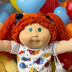 May include: A doll with bright orange yarn hair in pigtails, wearing a white shirt with a colorful fish pattern and white shoes. The doll is surrounded by a large yellow teddy bear, smaller dolls, heart-shaped balloons, and colorful candies.