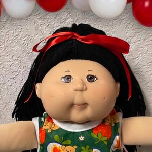 May include: A large doll with black yarn hair tied with a red ribbon, wearing a green sleeveless top featuring a strawberry and teddy bear print, white lace-trimmed socks, and surrounded by smaller dolls and red, white, and brown balloons.