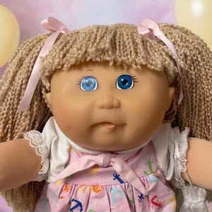 May include: A doll with yarn hair in pigtails, wearing a white shirt and a pink dress featuring a colorful anchor pattern. The doll has one blue eye and one light blue eye, and wears pink socks with ruffled trim. The doll is surrounded by smaller toys.