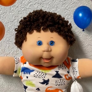 May include: A doll with brown curly hair and blue eyes wearing a white t-shirt with a dinosaur print and orange pants. The doll is surrounded by orange, black, blue, and white balloons, several colorful toy cars, and multiple Rubik's cubes.