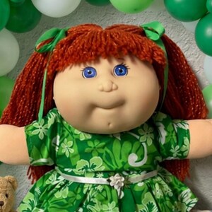 May include: A doll with red yarn hair and blue eyes wearing a green floral dress with a white belt, surrounded by a decorative arch of green and white balloons and four small brown plush teddy bears positioned around the doll's base.