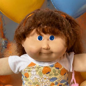 May include: A doll with brown hair and blue eyes wearing a white shirt with a yellow and orange strawberry print jumper, yellow ruffled socks, and white shoes, surrounded by orange, yellow, and blue teddy bears and heart-shaped balloons.