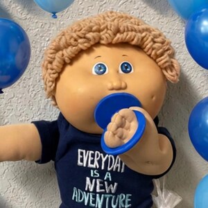 May include: A doll with tan skin, blue eyes, and curly light brown yarn hair, wearing a dark blue t-shirt with the text EVERYDAY IS A NEW ADVENTURE, dark blue jeans, and white shoes, with a blue pacifier, surrounded by blue balloons and toy cars.