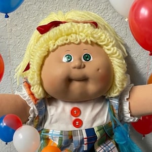 May include: A Cabbage Patch Kids doll with blonde yarn hair, green eyes, and a plaid dress with two red buttons, surrounded by smaller dolls, teddy bears, and colorful balloons. A blue Cabbage Patch Kids logo card sits on a patterned quilt.