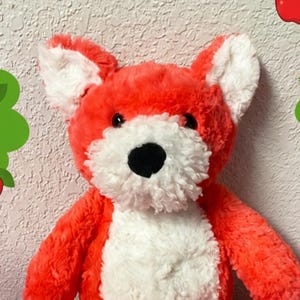 May include: A plush fox toy with bright orange fur, a white chest, white inner ears, and white paws. The fox has a black nose and dark eyes. It is positioned in the center of a digital frame featuring green trees, red apples, butterflies, ladybugs, and a caterpillar.