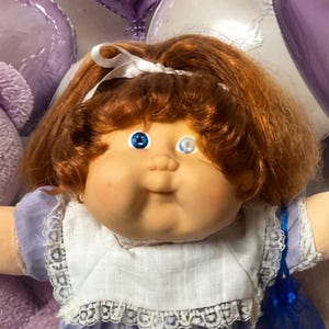 May include: A central doll with auburn hair and blue eyes wearing a light purple dress with white lace trim, flanked by two smaller dolls in pastel outfits, surrounded by purple heart balloons, brown brownies, and purple wrapped candies.