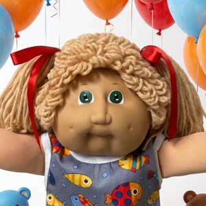 May include: A doll with yarn hair and red ribbons wearing a gray sleeveless dress with a colorful fish pattern, white socks, and white shoes. The doll is surrounded by small teddy bears in pink, blue, brown, and yellow, plus colorful candies and balloons.