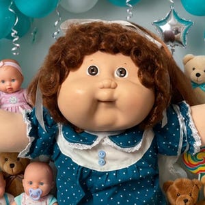 May include: A large doll with brown curly hair wearing a dark teal dress with white polka dots and a white collar, surrounded by several smaller baby dolls, brown teddy bears, colorful lollipops, chocolate chip cookies, and jelly beans.