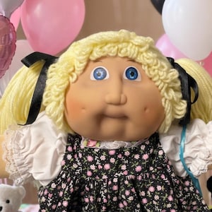May include: A doll with blonde yarn hair in pigtails, blue eyes, and dimpled cheeks, wearing a black dress with a small pink and white floral pattern, white lace trim, and white shoes. The doll is surrounded by small teddy bears and cupcakes.