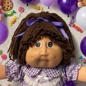 May include: A doll with brown yarn hair and a purple ribbon, wearing a purple and white gingham dress with a lace collar. The doll is surrounded by purple and white balloons, small teddy bears, baby dolls, chocolate chip cookies, and candies.