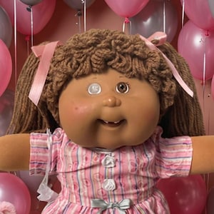 May include: A large doll with brown yarn hair and a pink striped dress sits surrounded by smaller dolls, two plush teddy bears, and scattered candy. The background is filled with pink and gray balloons, creating a festive display for the collection.