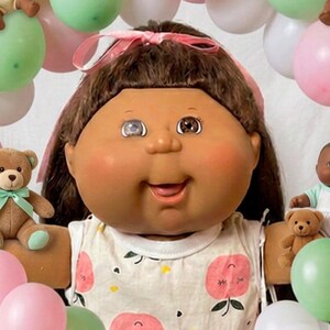 May include: A large doll with brown hair and a white sleeveless dress featuring a pink peach print, wearing white socks. The doll is surrounded by smaller dolls, miniature teddy bears, and a cluster of pink, white, and green balloons.