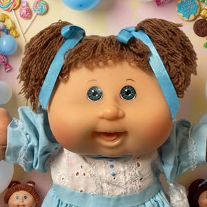 May include: A large doll with brown yarn hair in pigtails, wearing a light blue dress with white eyelet lace, surrounded by smaller dolls, blue and white balloons, assorted cookies, and colorful wrapped candies scattered throughout the scene.