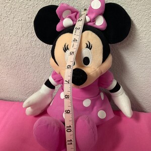 May include: A Minnie Mouse plush doll, approximately 30 cm tall, in a pink dress with white polka dots and a matching bow. The doll has black ears, a black nose, and white gloves. A measuring tape is placed next to the doll.
