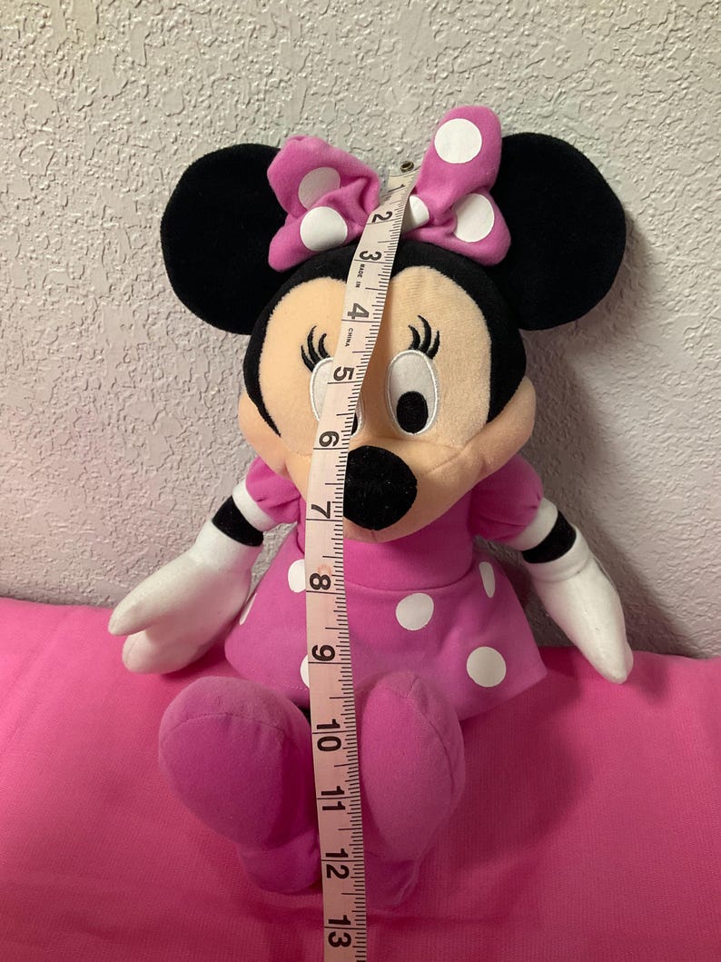 May include: A Minnie Mouse plush doll, approximately 30 cm tall, in a pink dress with white polka dots and a matching bow. The doll has black ears, a black nose, and white gloves. A measuring tape is placed next to the doll.