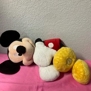 May include: A collection of plush Mickey Mouse items, including a head, two white gloves, a red and white sleeve, and two yellow plush items. The items are arranged on a pink surface.