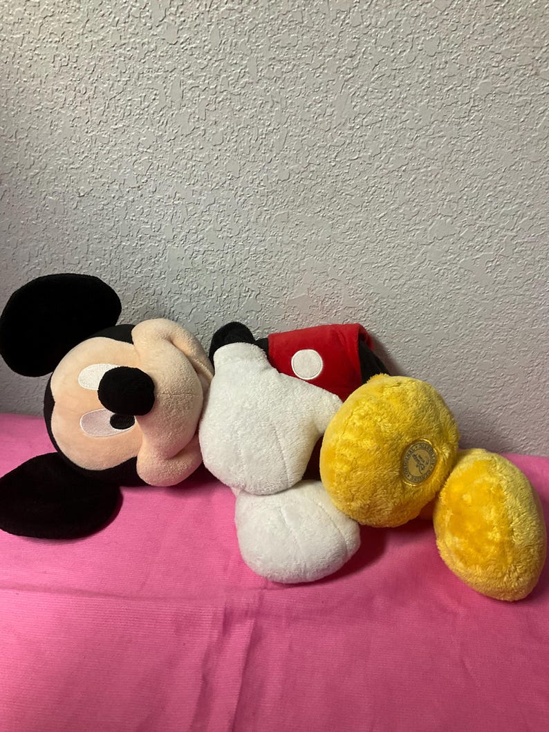May include: A collection of plush Mickey Mouse items, including a head, two white gloves, a red and white sleeve, and two yellow plush items. The items are arranged on a pink surface.