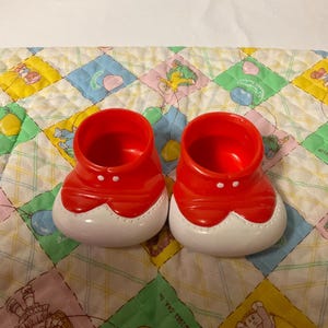 May include: A pair of red plastic doll shoes with white trim and two white buttons on each shoe.