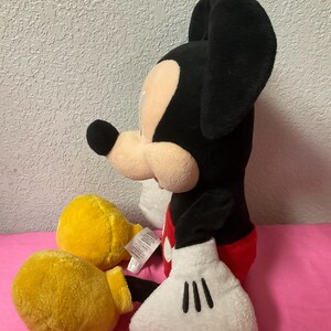 May include: A plush Mickey Mouse toy, featuring a black body, large black ears, and a tan face. It wears red shorts, yellow gloves, and white gloves with black details. The toy is sitting on a pink surface.
