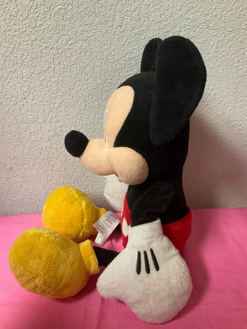 May include: A plush Mickey Mouse toy, featuring a black body, large black ears, and a tan face. It wears red shorts, yellow gloves, and white gloves with black details. The toy is sitting on a pink surface.