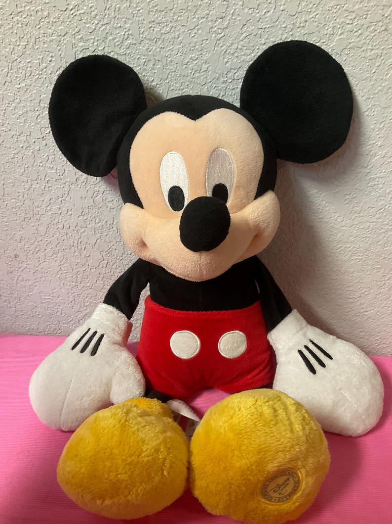 May include: A plush Mickey Mouse doll with black ears, a black nose, and a cream-colored face. The doll wears a red pair of shorts with two white buttons and white gloves. Two yellow plush balls are in front of the doll.