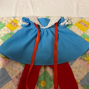 May include: A vintage doll outfit featuring a light blue dress with a white lace collar and red ribbons. The outfit includes red leggings. The ensemble is displayed on a colorful patchwork quilt.