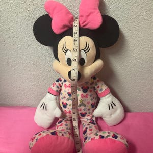 May include: A Minnie Mouse plush toy, approximately 21 inches tall, with a pink bow, black ears, and a patterned pajama set. The pajamas feature bows, stars, and rainbows. The toy has white gloves and pink feet.