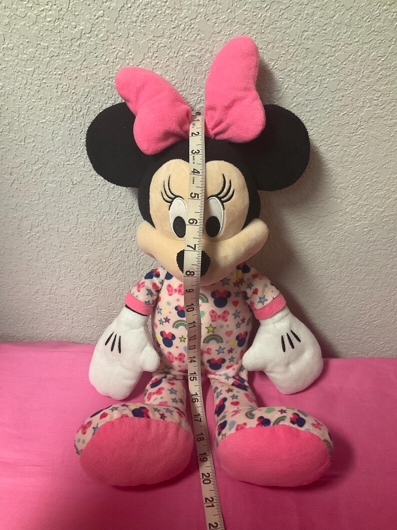May include: A Minnie Mouse plush toy, approximately 21 inches tall, with a pink bow, black ears, and a patterned pajama set. The pajamas feature bows, stars, and rainbows. The toy has white gloves and pink feet.
