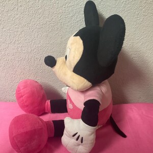 May include: A plush Mickey Mouse doll in a seated position. The doll has a black head and ears, a tan face, and a pink and white outfit. The doll's hands are white with black gloves, and its legs and feet are pink.