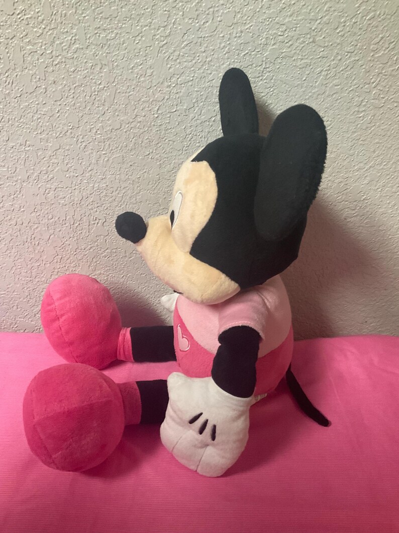 May include: A plush Mickey Mouse doll in a seated position. The doll has a black head and ears, a tan face, and a pink and white outfit. The doll's hands are white with black gloves, and its legs and feet are pink.