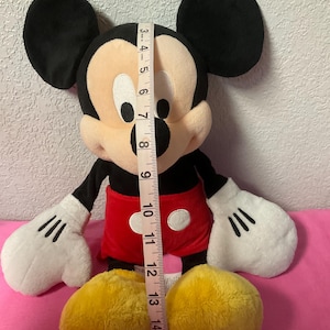 May include: A plush Mickey Mouse doll, approximately 12 inches tall, with black ears, a tan face, and a red shorts. The doll has white gloves and yellow shoes. A measuring tape is placed next to the doll.