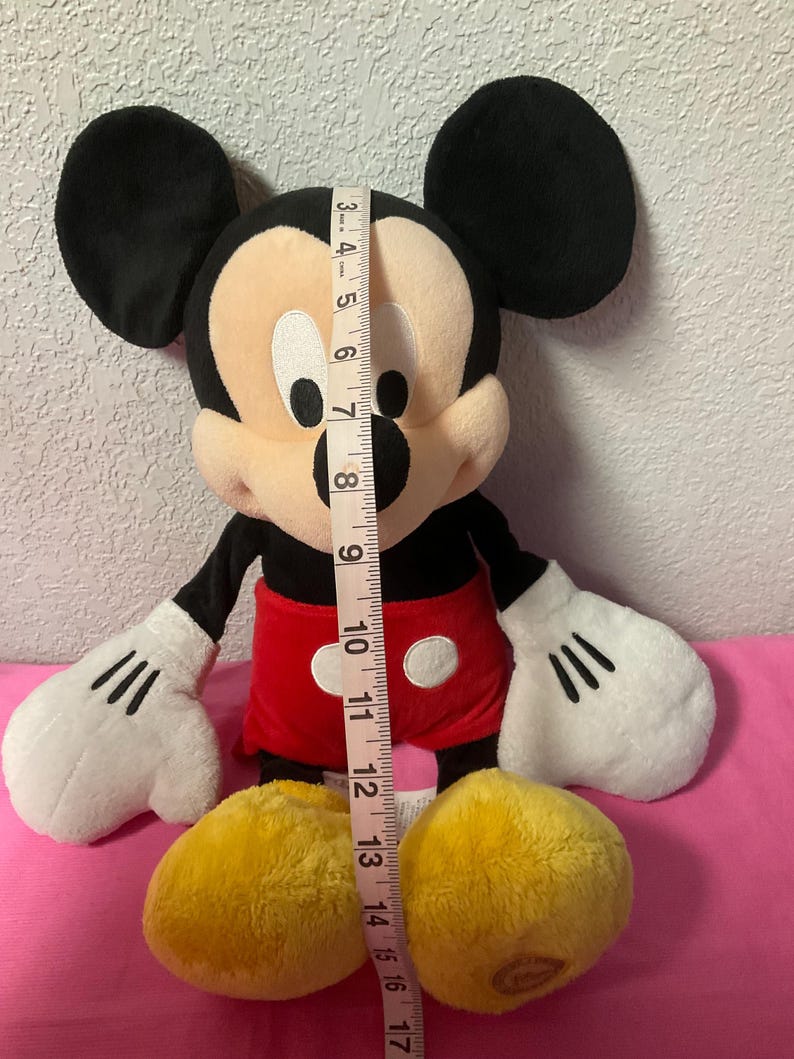 May include: A plush Mickey Mouse doll, approximately 12 inches tall, with black ears, a tan face, and a red shorts. The doll has white gloves and yellow shoes. A measuring tape is placed next to the doll.