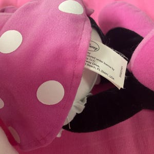 May include: A close-up of a pink plush Minnie Mouse bow with white polka dots. The bow is partially obscuring a black ear and a tag with the Disney logo and text. The background is a solid pink surface.