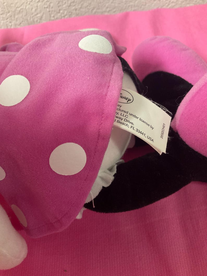 May include: A close-up of a pink plush Minnie Mouse bow with white polka dots. The bow is partially obscuring a black ear and a tag with the Disney logo and text. The background is a solid pink surface.