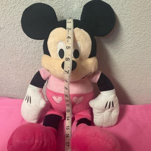 May include: A plush Mickey Mouse doll, primarily black, cream, and pink. The doll has large black ears, a cream-colored face, and wears a pink outfit with heart accents. The doll measures approximately 19 inches in height.