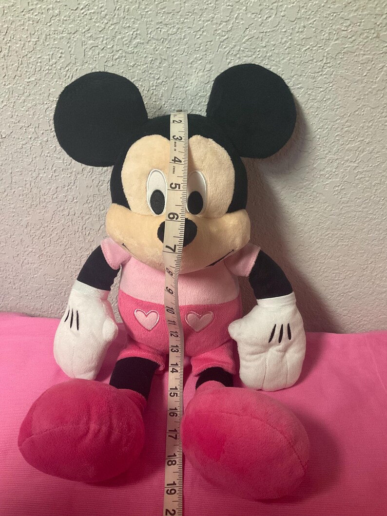 May include: A plush Mickey Mouse doll, primarily black, cream, and pink. The doll has large black ears, a cream-colored face, and wears a pink outfit with heart accents. The doll measures approximately 19 inches in height.