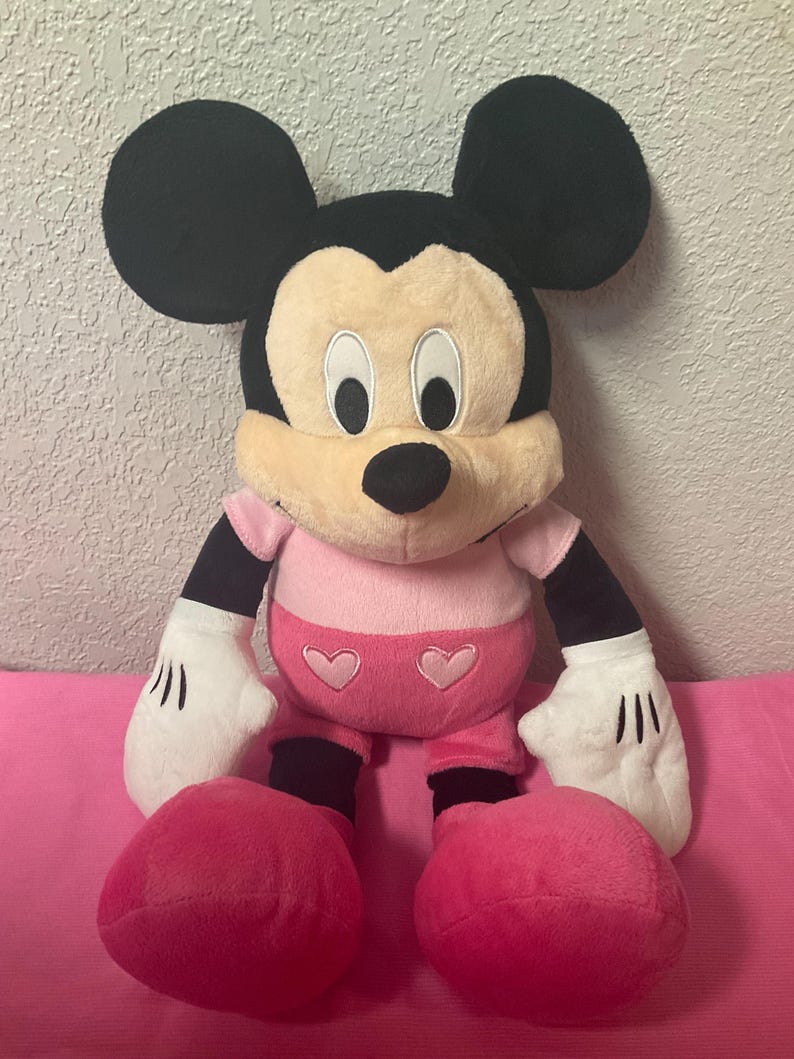 May include: A plush Mickey Mouse doll with black ears, a tan face, and a black nose. The doll wears a pink outfit with heart details and white gloves. The doll is sitting on a pink surface.