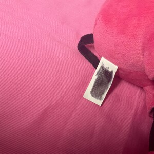 May include: A bright pink plush toy with a tag. The toy is partially visible, with a soft, textured surface. The tag is white with obscured text. The background is a matching pink fabric.