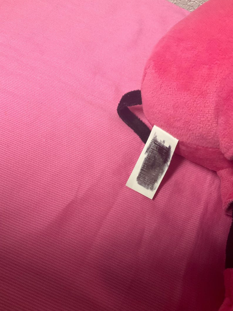 May include: A bright pink plush toy with a tag. The toy is partially visible, with a soft, textured surface. The tag is white with obscured text. The background is a matching pink fabric.
