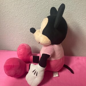 May include: A plush Mickey Mouse doll, featuring a black head with large ears, a cream-colored face, and a pink shirt and shorts. The doll has white gloves and large pink feet. The doll is sitting on a pink surface.