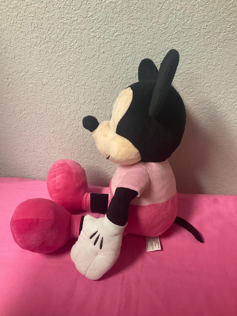 May include: A plush Mickey Mouse doll, featuring a black head with large ears, a cream-colored face, and a pink shirt and shorts. The doll has white gloves and large pink feet. The doll is sitting on a pink surface.