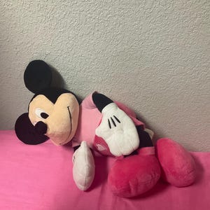 May include: A plush Minnie Mouse doll, featuring a black head with large ears, a tan face, and a pink dress. The doll has white gloves and pink shoes. The doll is lying on a pink surface.