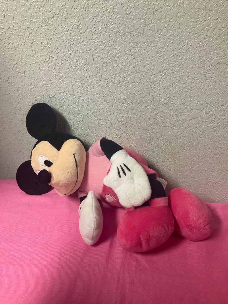 May include: A plush Minnie Mouse doll, featuring a black head with large ears, a tan face, and a pink dress. The doll has white gloves and pink shoes. The doll is lying on a pink surface.