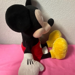 May include: A plush Mickey Mouse toy, featuring a black head and ears, a white face, and a black nose. The toy wears red shorts, white gloves, and yellow shoes. The stuffed animal is sitting on a pink surface.
